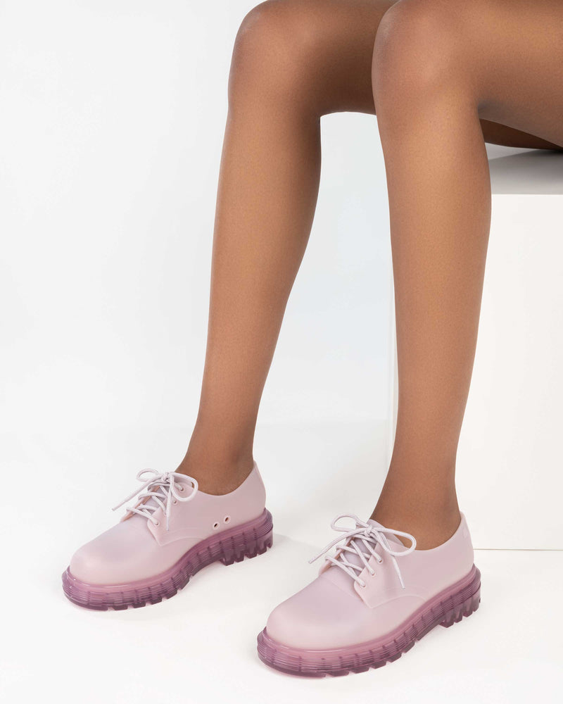 A model's legs wearing a pair of matte lilac Melissa Bass sneakers, with a light matte lilac base, lilac laces and a darker transparent lilac sole.