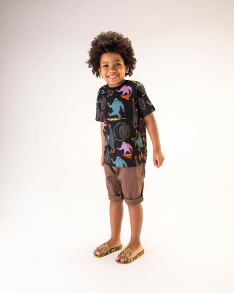 A model wearing a black patterned t-shirt, brown shorts and a pair of clear beige / yellow Mini Melissa Beach slides with a beige base and a pattern of Disney's Pluto the dog with different facial expressions.