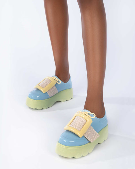A model's legs wearing a pair of blue Melissa Buckle Up Kick Off platform sneaker with green platform sole and a yellow buckle and ivory strap.