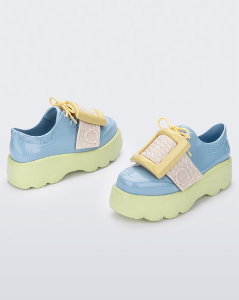 Angled view of a pair of blue Melissa Buckle Up Kick Off platform sneaker with green platform sole and a yellow buckle and ivory strap.