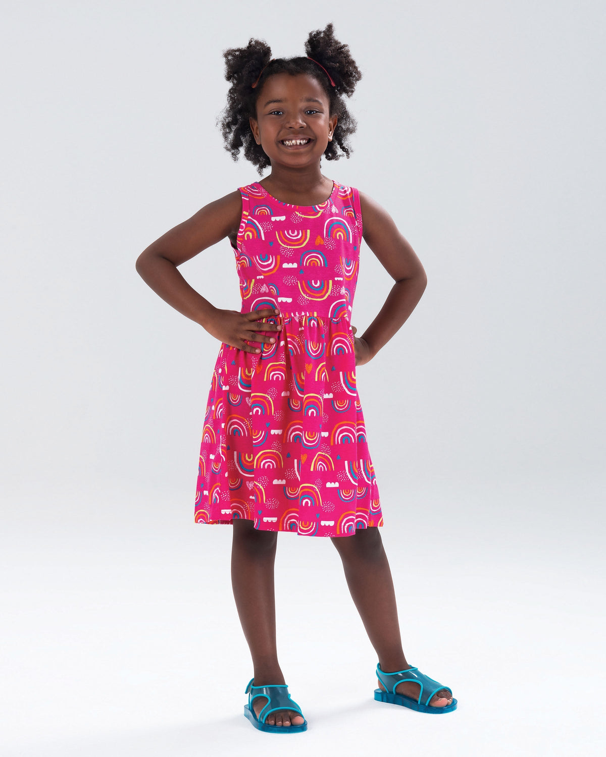 A model posing in a pink patterned dress wearing the Mini Melissa Aqua sandals in blue, with two straps cojoining in the middle and a back strap with velcro closure.