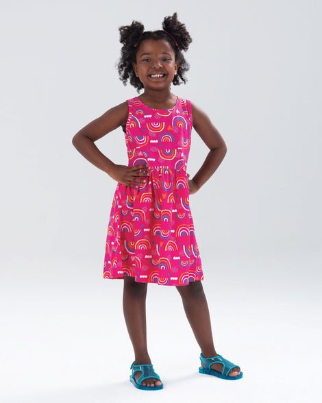 A model posing in a pink patterned dress wearing the Mini Melissa Aqua sandals in blue, with two straps cojoining in the middle and a back strap with velcro closure.