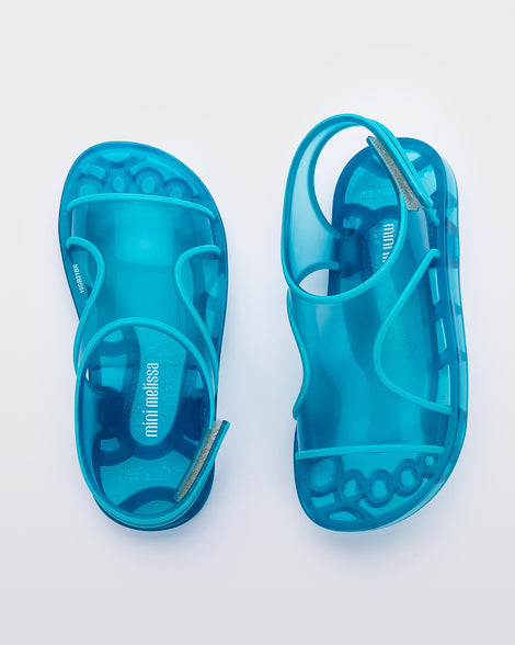 A top front and back view of a pair of translucent blue Mini Melissa Aqua sandals with two straps cojoining in the middle and a velcro back strap.