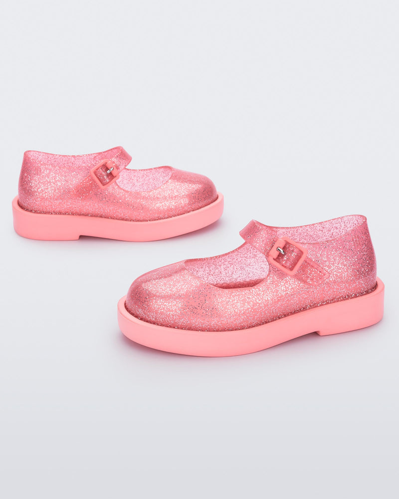 A side view of a pair of transparent glitter pink Mini Melissa Lola loafer with a rounded toe and a pink strap and buckle.