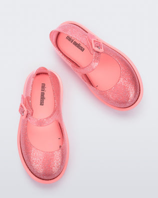 A top view of a pair of transparent glitter pink Mini Melissa Lola loafer with a rounded toe and a pink strap and buckle.