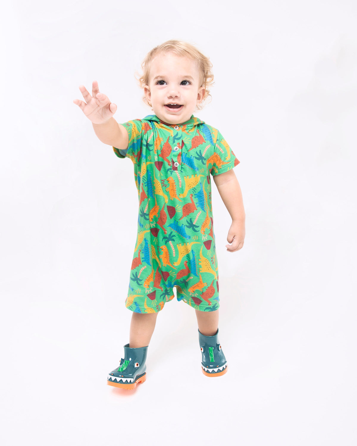 A baby model posing for a picture in a dinosaur onesie and a pair of orange/green Mini Melissa Rain Boots with a green base, orange sole, light green triangle details, white triangle details in the front and a set of orange eyes which looks like a dinosaurs face,