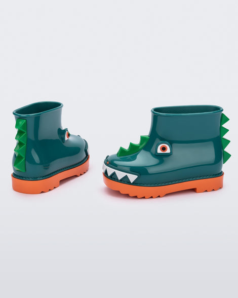 An angled side and back view of a pair of orange/green Mini Melissa Rain Boots with a green base, orange sole, light green triangle details, white triangle details in the front and a set of orange eyes which looks like a dinosaurs face,