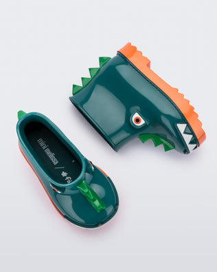 A top and side view of a pair of orange/green Mini Melissa Rain Boots with a green base, orange sole, light green triangle details, white triangle details in the front and a set of orange eyes which looks like a dinosaurs face,