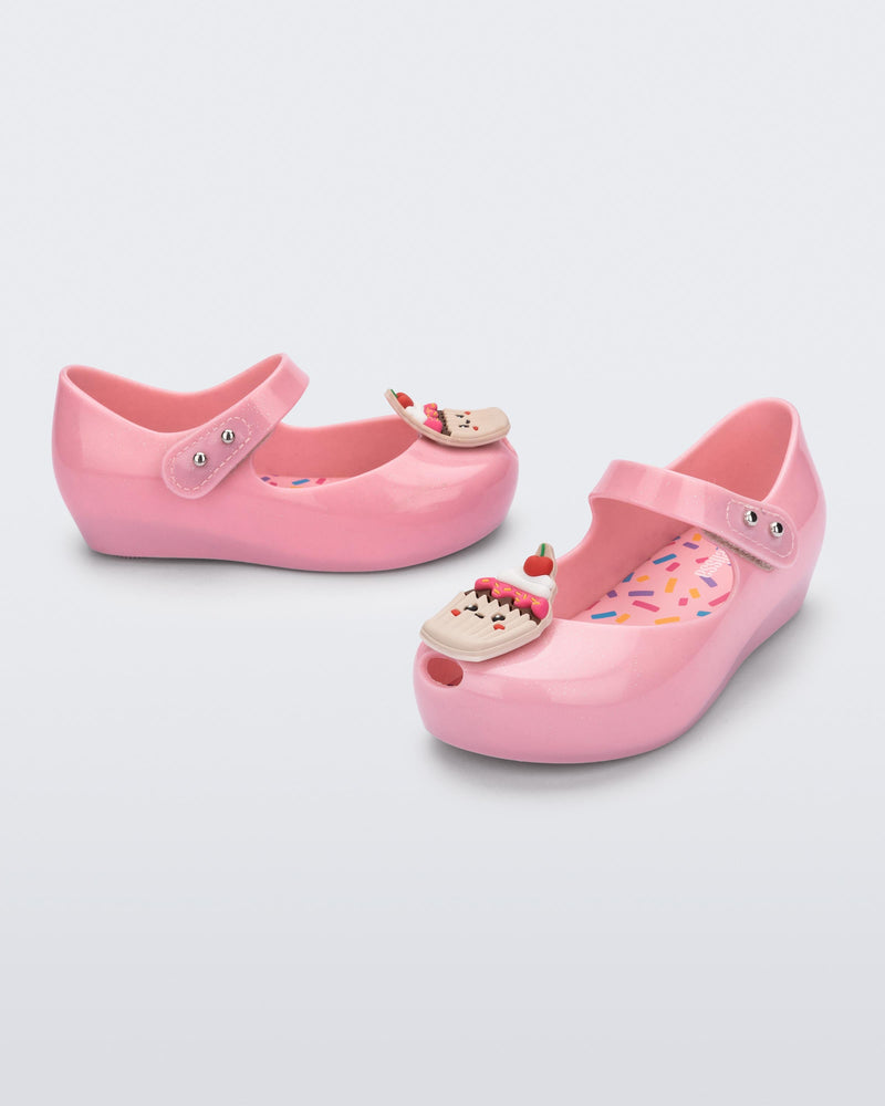 Angled view of a pair of Mini Melissa Ultra Girl Candy ballet flats in pink glitter colorway. The shoe has a single strap on the top of the foot with a double snap closure. The shoe has a 3D printed cupcake applicque with smiling face on the top of the toe. The colors of the applique are red, brown and white. 