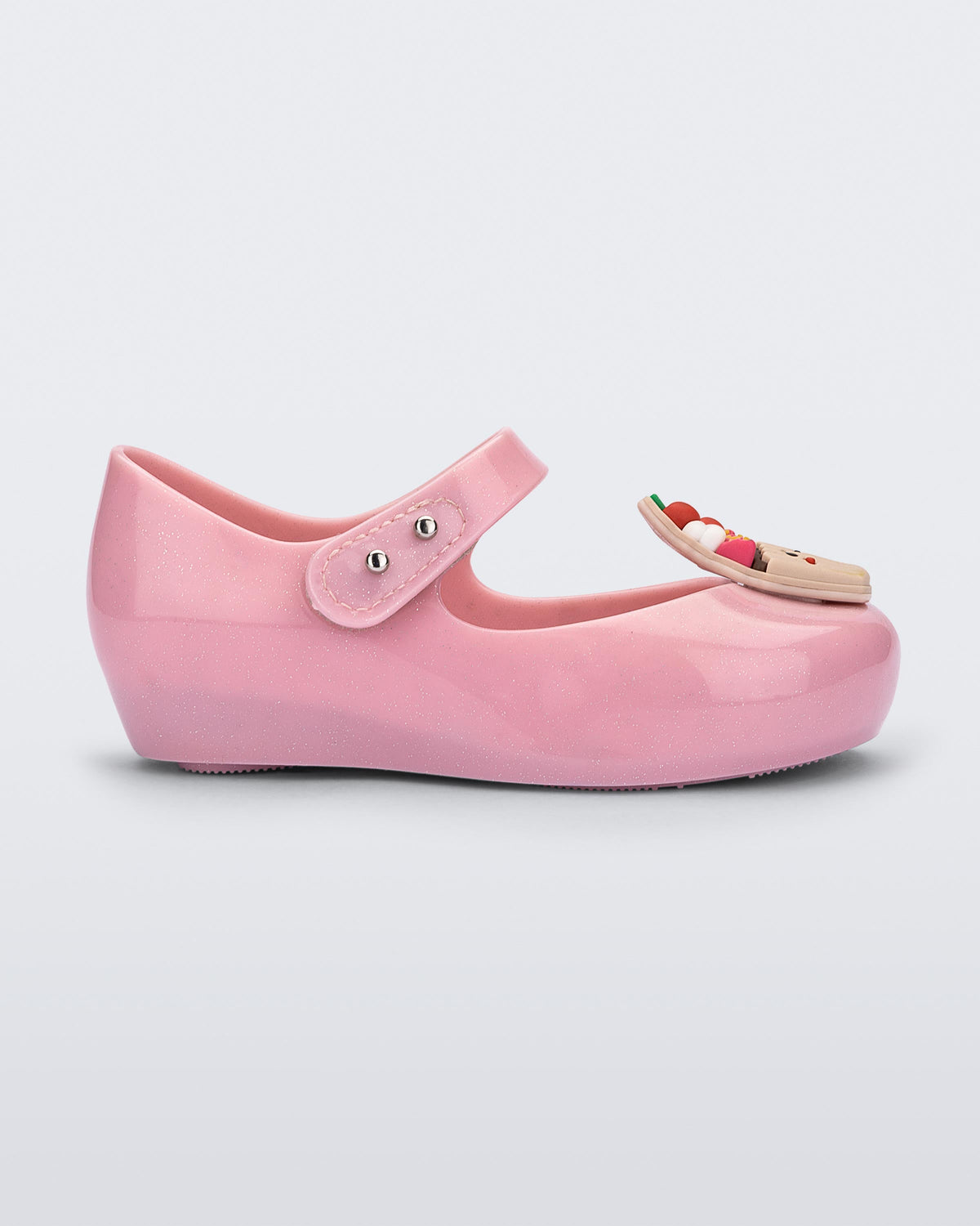 Side view of a Mini Melissa Ultra Girl Candy ballet flat in pink glitter colorway. The shoe has a single strap on the top of the foot with a double snap closure. The shoe has a 3D printed cupcake applicque with smiling face on the top of the toe. The colors of the applique are red, brown and white. 