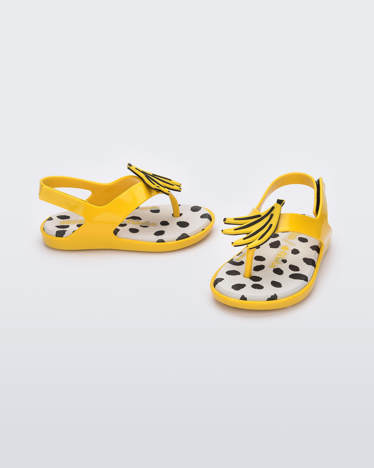 An angled front and side view of a pair of yellow Mini Melissa Sunny sandals with a black base, a banana drawing on top of the straps and a black and white polka dot insole.