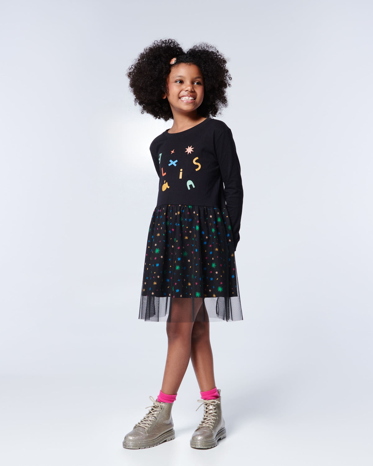 Model posing in a black and rainbow print dress wearing a pair of Mini Melissa lace up Coturno boots in gold glitter color-way paired with bright pink ankle socks.