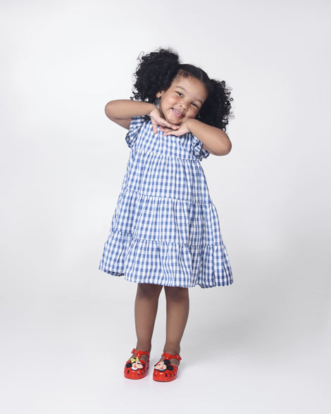 Girl in a white and blue dress wearing a pair of red Mini Melissa Possession + Disney 100 sandals with Mickey on one foot and Minnie on another foot.