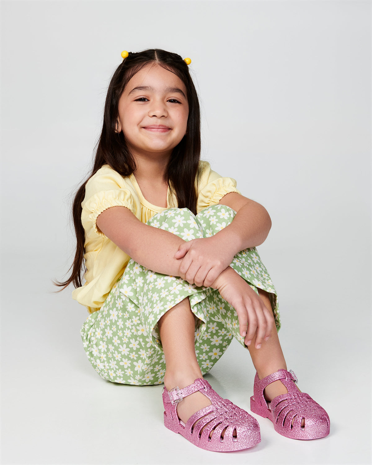 Child model in a yellow shirt and green pants wearing a pair of glitter pink kids Possession sandals.