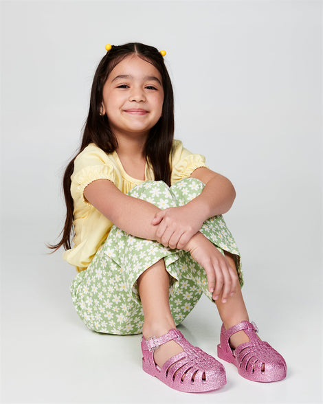Child model in a yellow shirt and green pants wearing a pair of glitter pink kids Possession sandals.