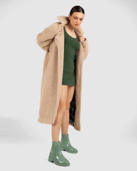 Model wearing a green dress and brown long jacket wearing a pair of sage green Chelsea Boots.
