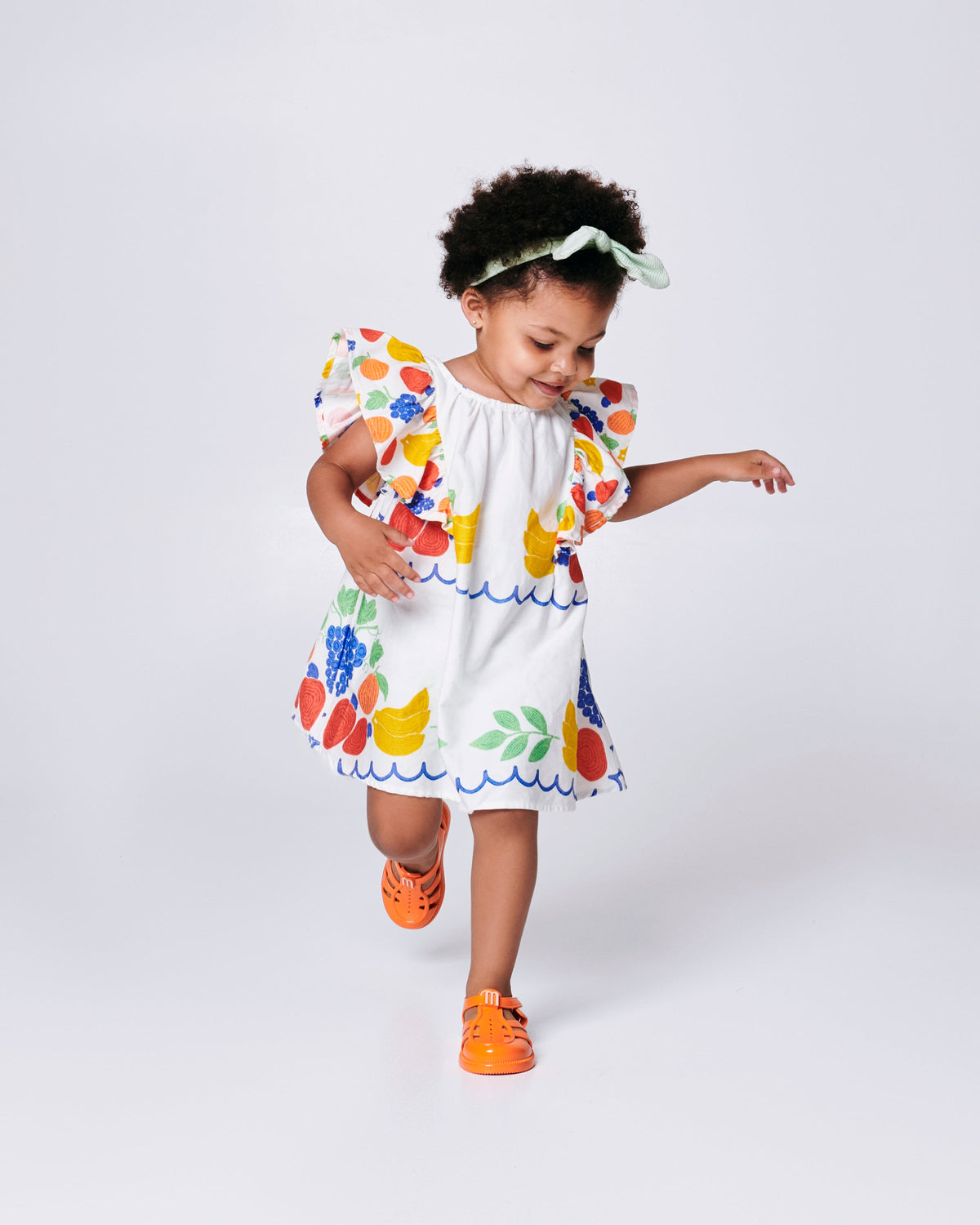 Child model in a white dress with flower print wearing a pair of orange Hip Daydream baby sandals