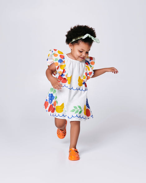 Child model in a white dress with flower print wearing a pair of orange Hip Daydream baby sandals