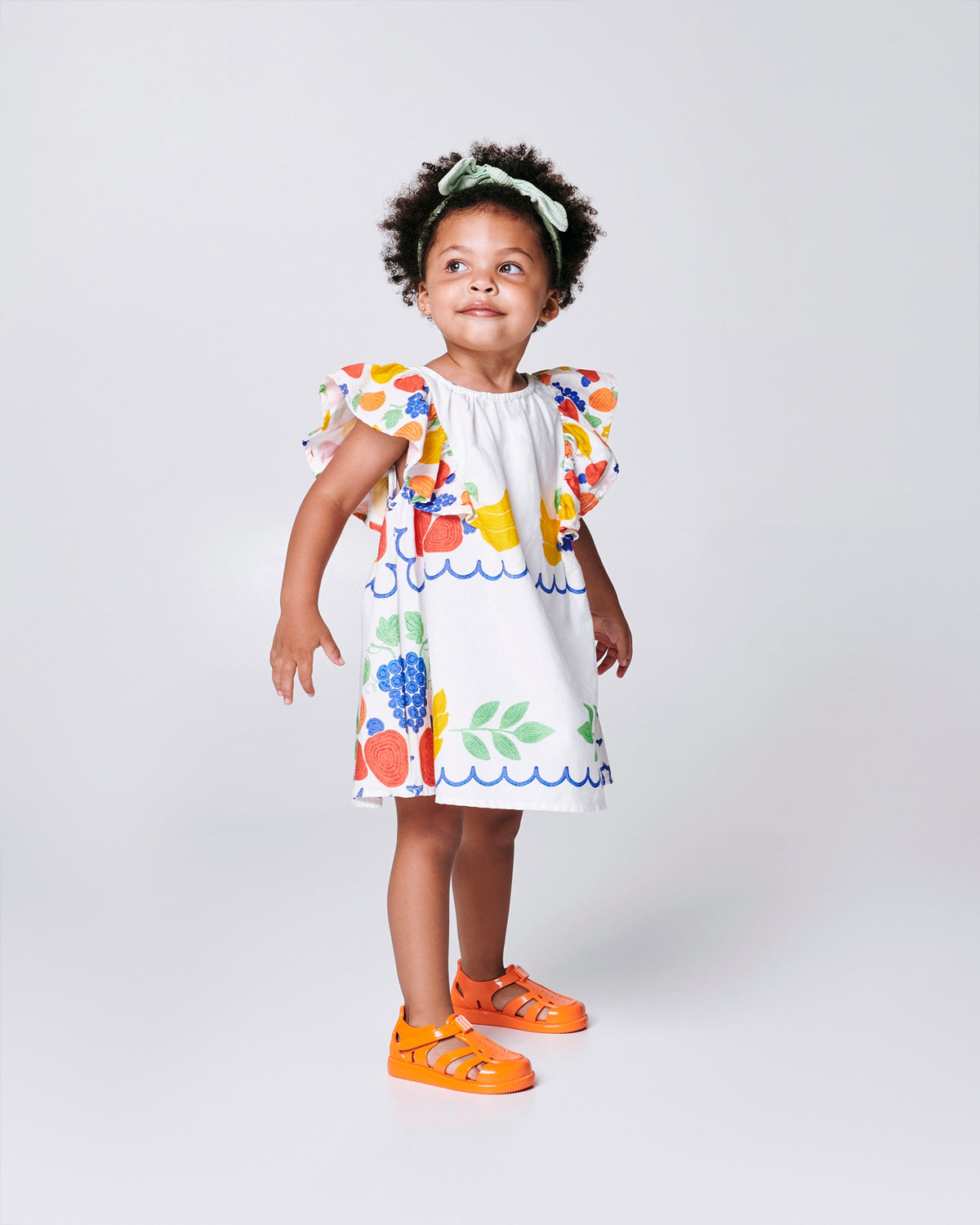Child model in a white dress with flower print wearing a pair of orange Hip Daydream baby sandals