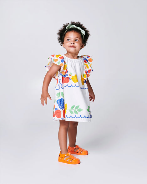 Child model in a white dress with flower print wearing a pair of orange Hip Daydream baby sandals
