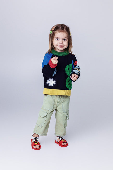Child model in a sweater and green pants wearing a pair of red  Play Mickey & Minnie sandals with white straps