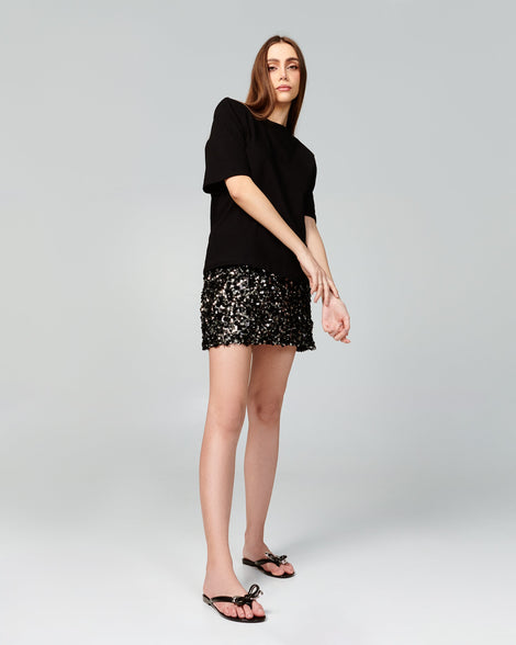 Model in a black sweater and sparkling skirt wearing a pair of black Harmonic Eclipse flip flops with bow.