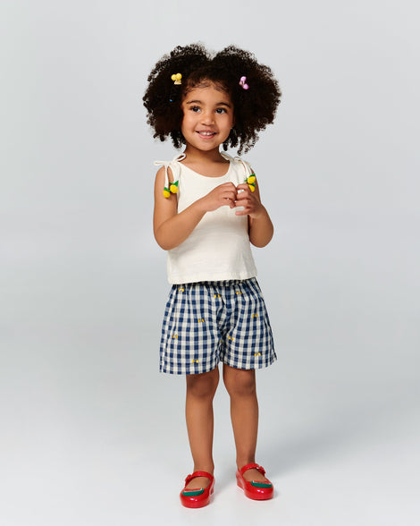 Child model in a white shirt and black and white shorts wearing a pair of red Sweet Love Picnic baby flats with watermelon slices