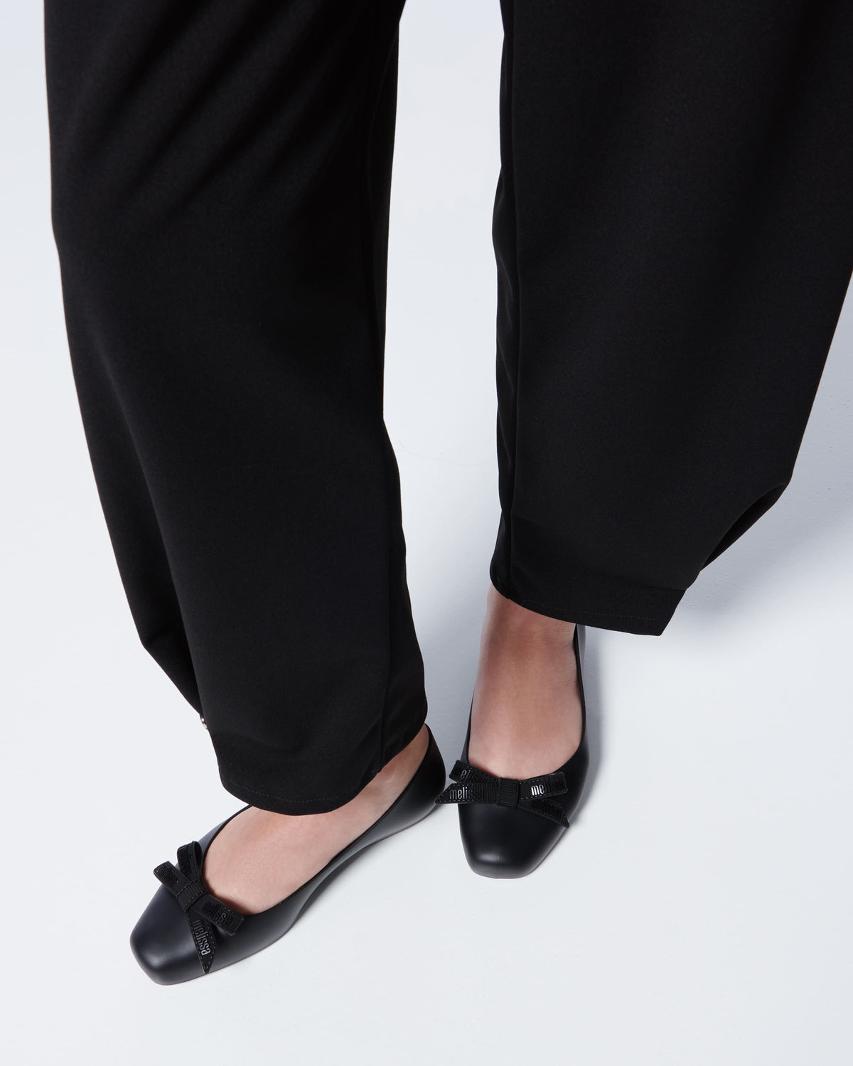 Close up of a model's legs wearing lack pants and Melissa Aura flats in black with a bow on a white background