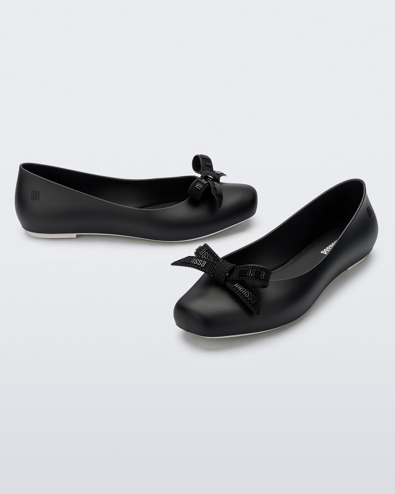 Angled view of a pair of Melissa Aura flats in black  with bow details on a light gray background