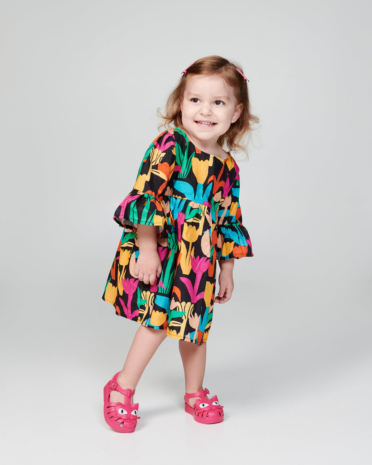 Child model in a black print dress wearing a pair of pink baby Possession Cat sandals