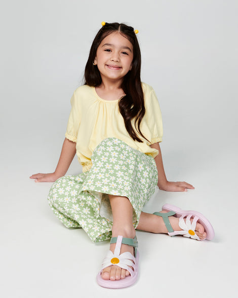 Child model in a yellow shirt and green floral pants wearing a pair of pink and green kids Hip Bloom sandal.