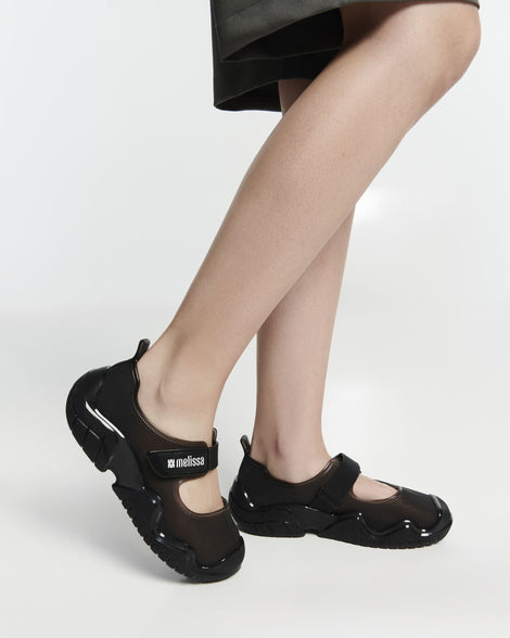 Close up view a model's legs in a pair of the Melissa Sprint Ballerina flats in black  with velcro straps on a light gray background