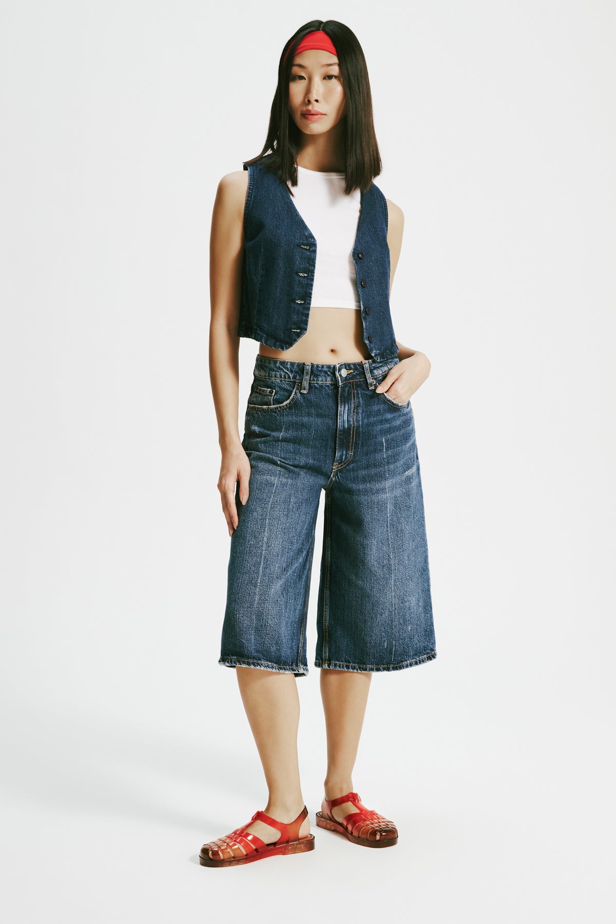 Model in a white shirt with a jean vest and jean shorts wearing a pair of transparent red Possession + Nodaleto fisherman sandals.