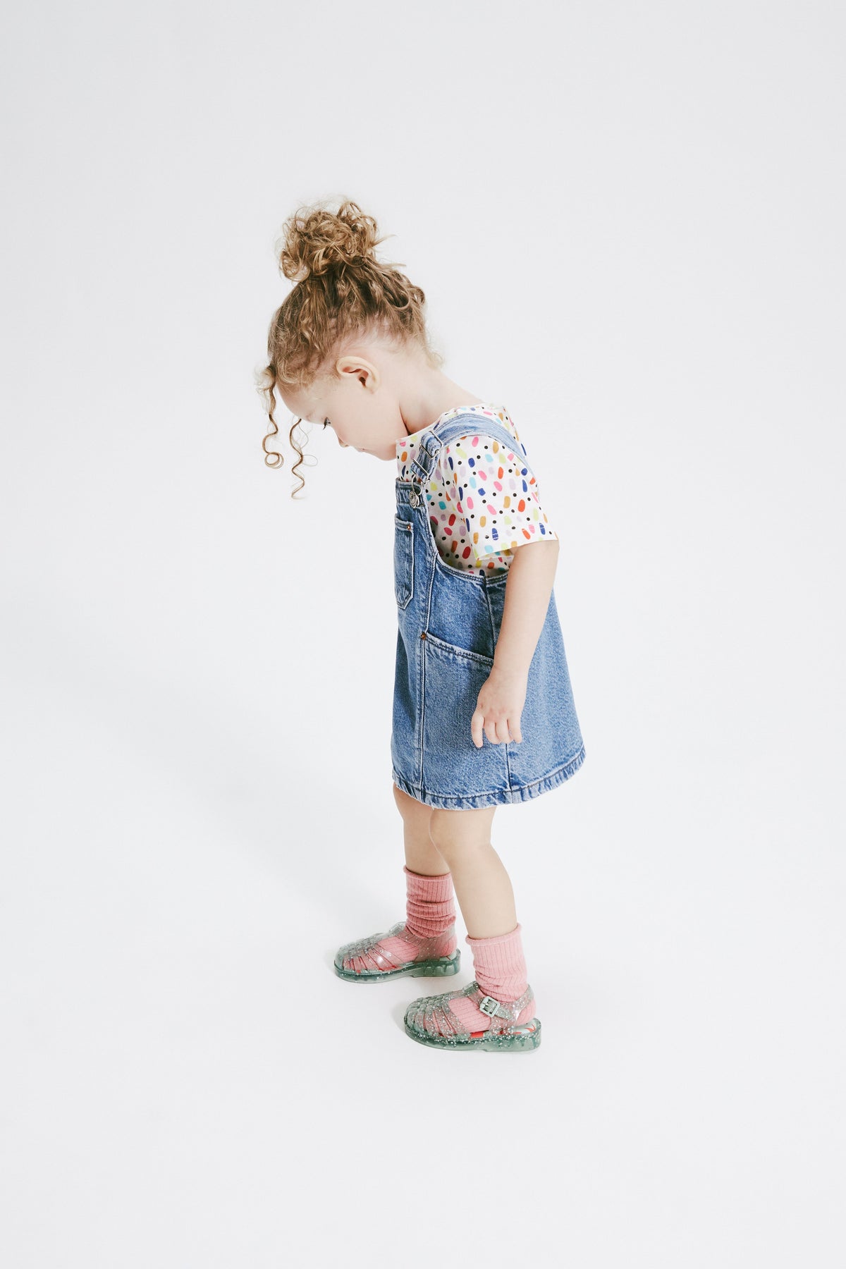 Child model in a jean overall dress wearing glitter green Possession baby sandals with orange socks