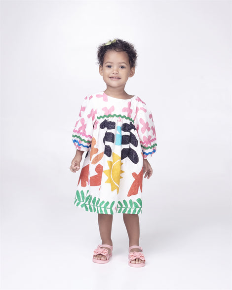 A baby model posing for a picture in a white patterned dress and a pair of glitter pink Mini Melissa Mar Sandal Heart sandals with a glitter pink heart bow detail on the front strap and an ankle strap