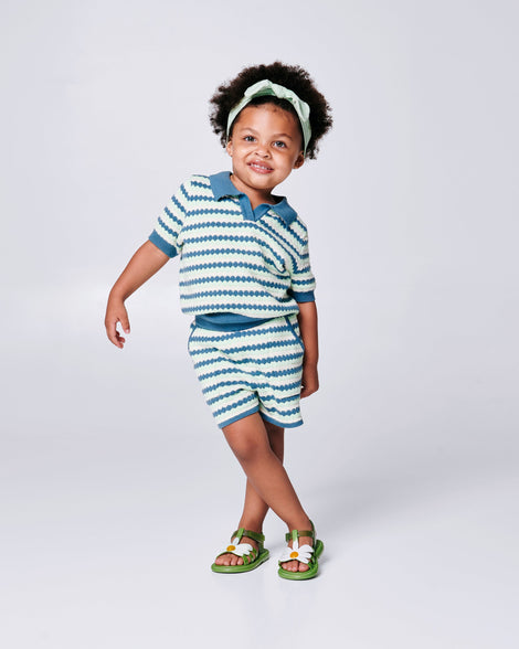 Child model in a blue and white outfit wearing a pair of green baby Hip Bloomy sandal with a white flower