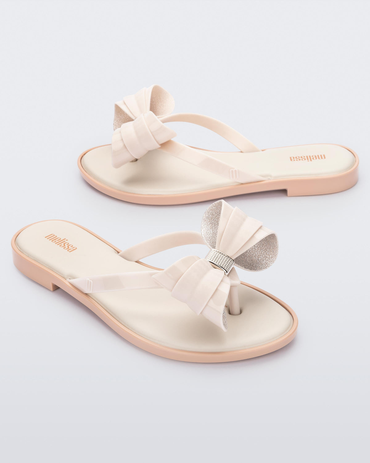 Angled view of a pair of Melissa slim strap flip flops in light beige with bow applique