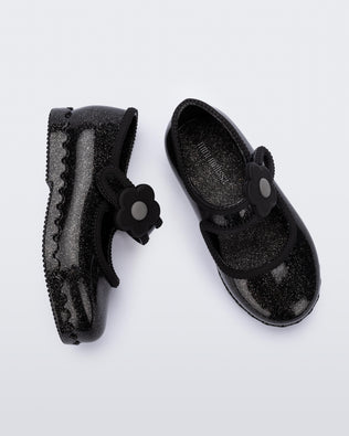 Side and top view of a pair of black Hip Ballerina baby flats with strap with flower