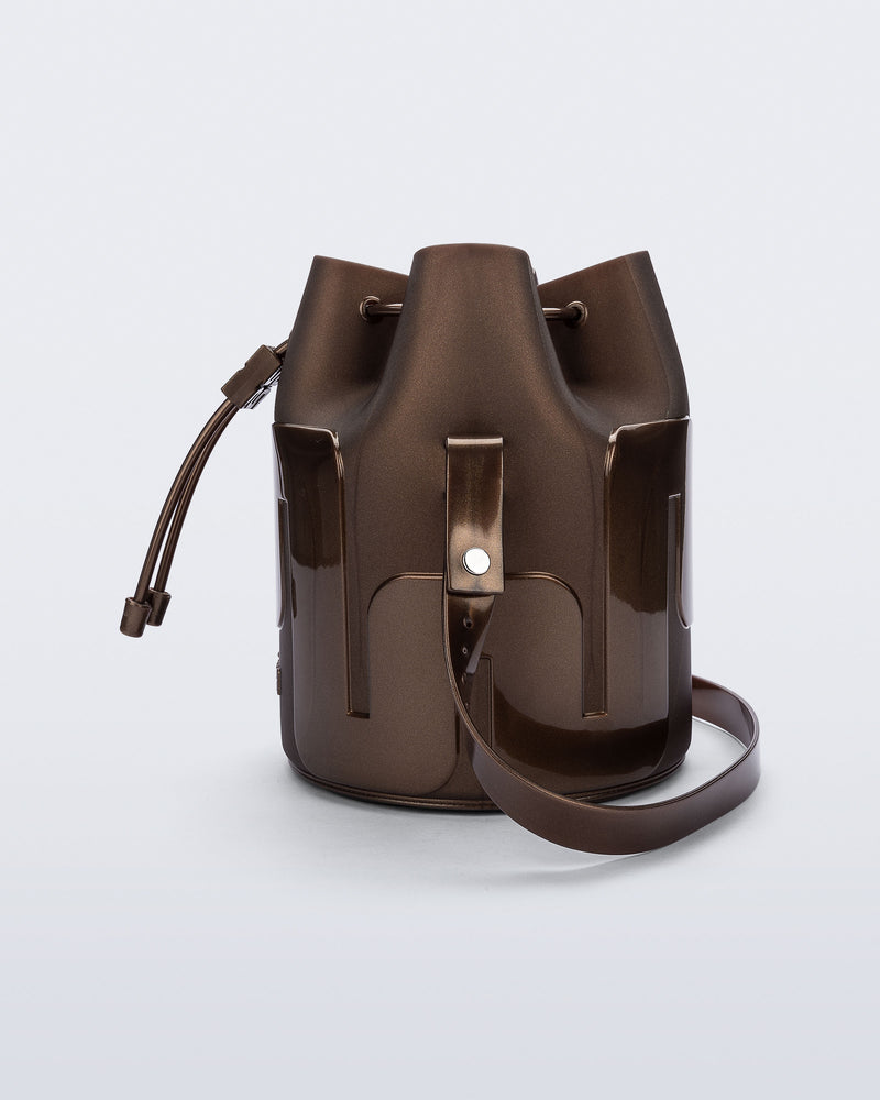 Side view of a Melissa Station bucket bag in Bronze. The bag has an adjustable shoulder strap and a drawstring top closure. 