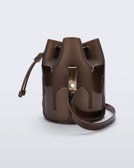 Side view of a Melissa Station bucket bag in Bronze. The bag has an adjustable shoulder strap and a drawstring top closure. 