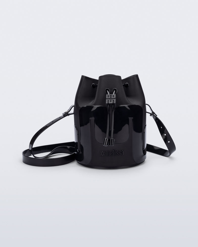 Side view of a Melissa Station bucket bag in Black. The bag has an adjustable shoulder strap and a drawstring top closure. The Melissa logo is on the bottom center of the front of the bag. 