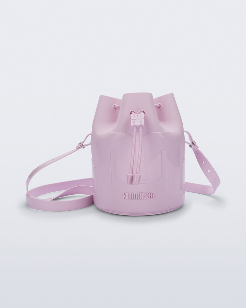 Side view of a Melissa Station bucket bag in Light Pink. The bag has an adjustable shoulder strap and a drawstring top closure. The Melissa logo is on the bottom center of the front of the bag. 