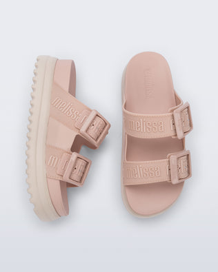 Top and side view of a pair of melissa cozy platform slide sandals. There are two buckle straps that go across the foot with the M Lover (melissa logo) print embossed into the straps. The buckles are located on the outside edge of the two straps. The straps and insole of the shoe are light pink and the platform sole is beige. 