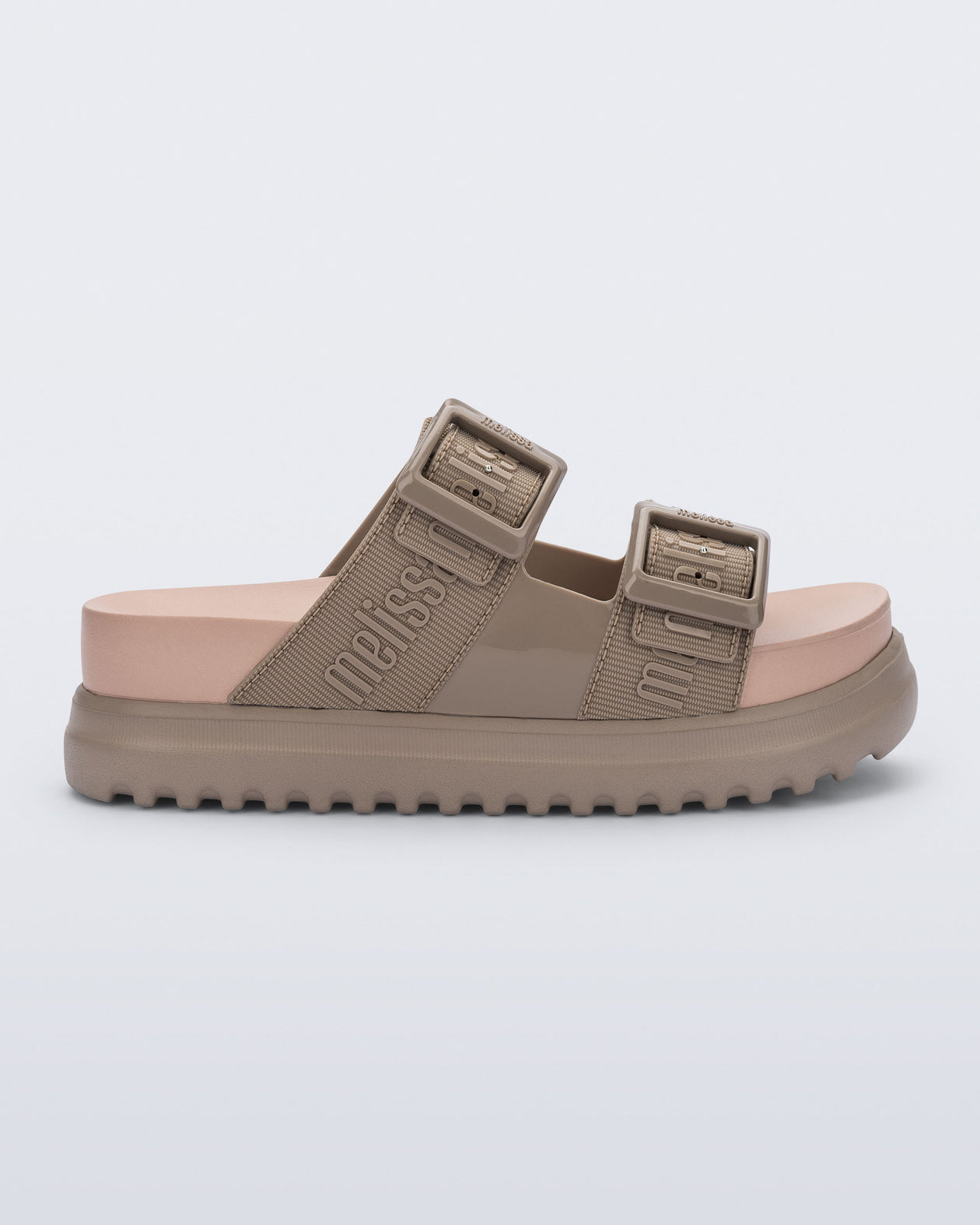 Side view of the melissa cozy platform slide sandal. There are two buckle straps that go across the foot with the M Lover (melissa logo) print embossed into the straps. The buckles are located on the outside edge of the two straps. The straps and platform sole of the shoe are dark beige color and the insole is a lighter color beige. 