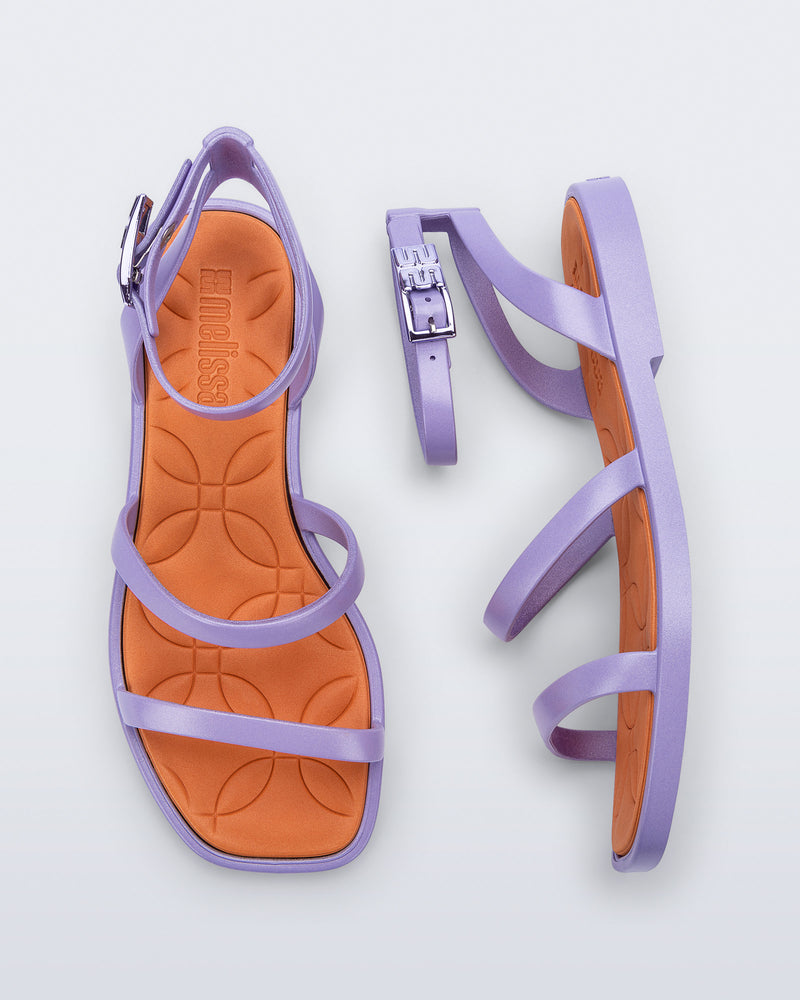 Top and side view of a pair of Melissa Heat Sandals. This is a flat sandal with a multiple straps, one across the toes, mid part of the foot, and a buckle strap around the ankle with a supporting strap that connects the ankle strap to the sole of the shoe. The straps and outsole are purple and the insole of the shoe is orange. The insole is embossed with a geometric pattern. 