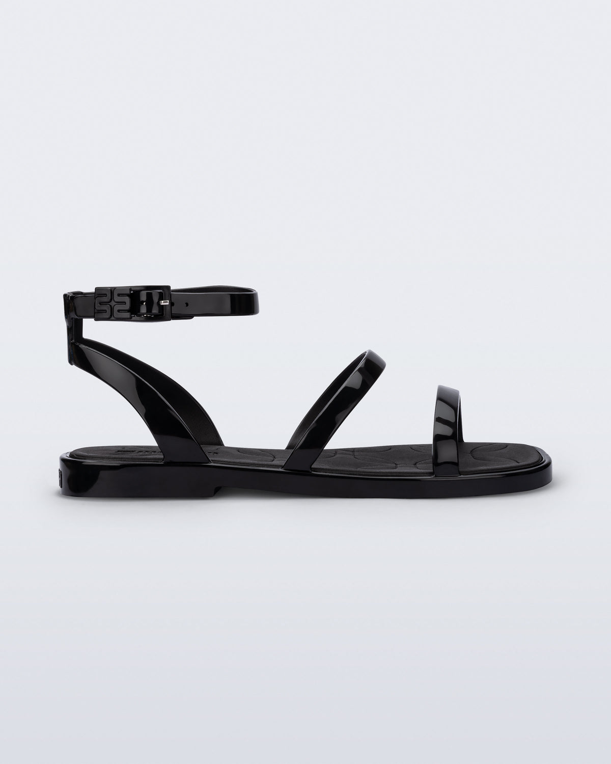 Side view of the Melissa Heat Sandal. This is a flat sandal with a multiple straps, one across the toes, mid part of the foot, and a buckle strap around the ankle with a supporting strap that connects the ankle strap to the sole of the shoe. The straps, insole, outsole, and buckles on the ankle strap are black. The insole is embossed with a geometric pattern. 