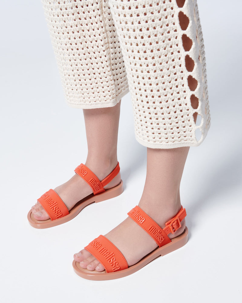 Close up view of a model's legs posing in a pair of Melissa Mar Sandals with Melissa logo embossed into the straps. There are two embossed straps that go across the foot; 1 across the toes and the other across the main part of the foot. The main foot strap has an ankle strap attached with a buckle closure. The straps and buckle are orange and the sole of the shoe is brown. The buckle placement is on the outside of the ankle strap. 