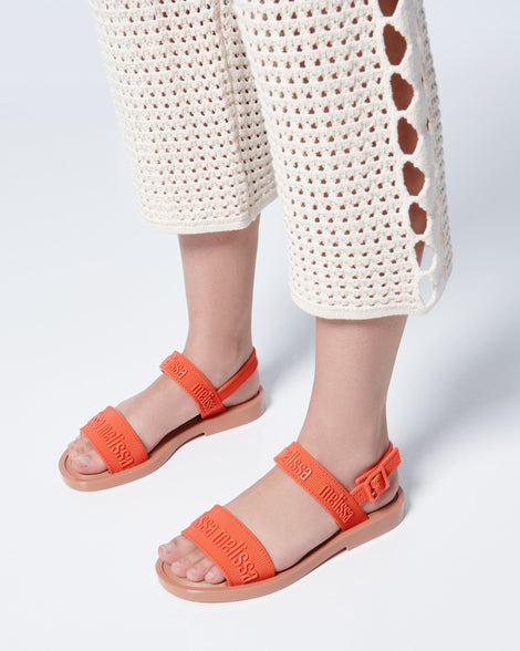 Close up view of a model's legs posing in a pair of Melissa Mar Sandals with Melissa logo embossed into the straps. There are two embossed straps that go across the foot; 1 across the toes and the other across the main part of the foot. The main foot strap has an ankle strap attached with a buckle closure. The straps and buckle are orange and the sole of the shoe is brown. The buckle placement is on the outside of the ankle strap. 