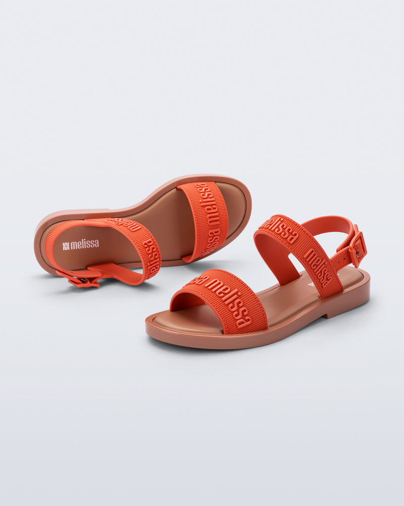 Top and side view of a pair of Melissa Mar Sandals with Melissa logo embossed into the straps. There are two embossed straps that go across the foot; 1 across the toes and the other across the main part of the foot. The main foot strap has an ankle strap attached with a buckle closure. The straps and buckle are orange and the sole of the shoe is brown. The buckle placement is on the outside of the ankle strap. 