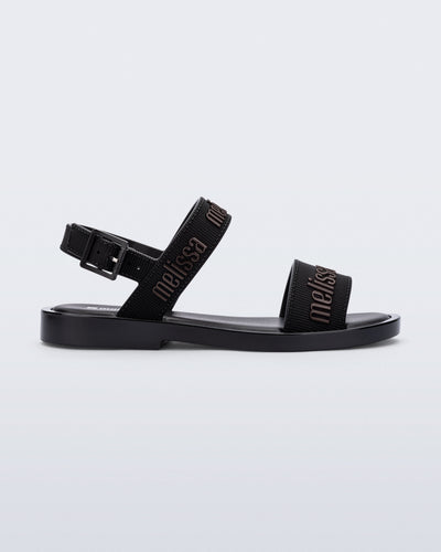 Side view of the Melissa Mar Sandal with Melissa logo embossed into the straps. There are two embossed straps that go across the foot; 1 across the toes and the other across the main part of the foot. The main foot strap has an ankle strap attached with a buckle closure. The straps ,buckle and sole of the shoe is black. The Melissa logo on the straps has a black metallic finish. . The buckle placement is on the outside of the ankle strap. 